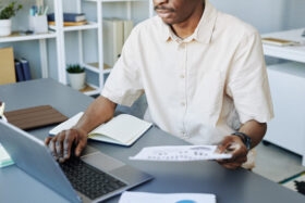 Cropped shot of adult African American man reading data reports while working in office and using laptop.